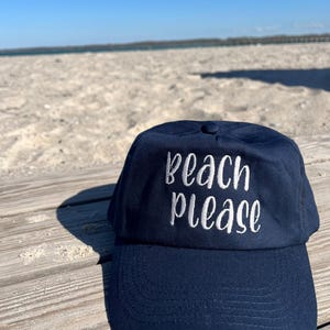 Può includere: Un cappellino da baseball blu navy con la scritta "beach please" ricamata in bianco sul pannello frontale. Il cappellino presenta una corona strutturata, una visiera curva con cuciture visibili e un bottone al centro della calotta.