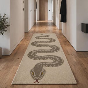 May include: A rectangular runner rug featuring a large, winding snake motif in shades of grey and brown. The snake has a textured pattern, yellow eyes, and a red forked tongue, set against a light beige background, positioned on a wooden floor.