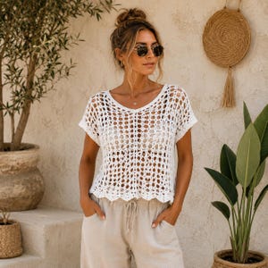 Può includere: Un top a maniche corte in crochet bianco con motivo a maglia aperta e orlo smerlato, abbinato a pantaloni beige chiaro con coulisse. Il capo presenta un'ampia scollatura e una vestibilità morbida, caratterizzata da una texture artigianale.