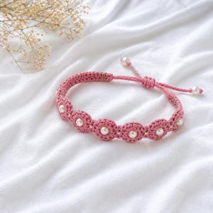 Puede incluir: Una pulsera de ganchillo rosa con detalles de perlas blancas. La pulsera presenta una serie de diseños en forma de flor de ganchillo, cada uno centrado con una perla. El cierre ajustable es de cordón rosa y tiene una perla en cada extremo. La pulsera se muestra sobre una superficie blanca.