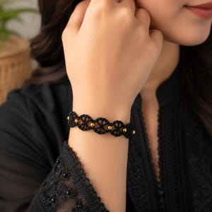 Könnte beinhalten: Ein schwarzes Armband aus geflochtener Kordel mit einem sich wiederholenden kreisförmigen Muster und kleinen, polierten goldfarbenen Kugeln, die zwischen jedem geflochtenen Glied eingearbeitet sind und ein durchgehendes, symmetrisches Design bilden.