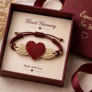 May include: A handmade macrame bracelet with a red heart and beige wings, presented in a burgundy gift box. The box has the text "Heart Harmony" and "Made with love". The gift box also has the text "Especially For You".