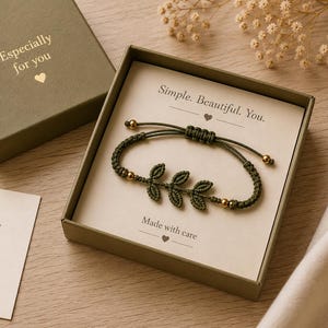 Puede incluir: Una pulsera de macramé verde oliva con diseño de hojas y cuentas doradas, presentada en una caja de regalo. La caja es verde oliva y tiene el texto "Especially for you". La tarjeta dentro de la caja dice "Simple. Beautiful. You."