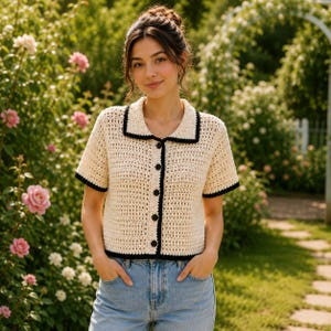 May include: A short-sleeved, cream-coloured crochet shirt featuring a black collar, black sleeve trim, and a black front placket with five dark buttons. The garment has a textured, open-knit pattern and is worn with light blue denim jeans.