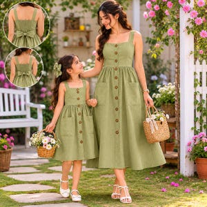 May include: Two olive green dresses with button-down fronts and square necklines. The dresses have pockets and are worn by a woman and a girl. The girl carries a basket of flowers. The dresses are a matching set.