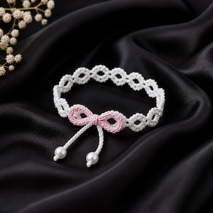 May include: A white crocheted choker necklace with a pink bow and two white pearl beads. The necklace is set against a black satin background. The choker has a delicate, openwork design.
