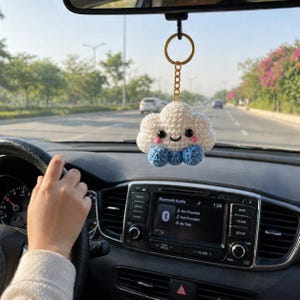 Puede incluir: Un adorno de coche de nube hecho a mano con un rostro sonriente, detalles azules y un llavero dorado. El adorno cuelga del espejo retrovisor de un coche. Se ve el salpicadero del coche y una mano en el volante.