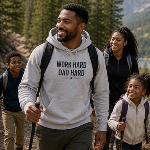 May include: A light gray hooded sweatshirt featuring the black text WORK HARD DAD HARD printed on the chest. The garment includes a front kangaroo pocket, long sleeves with ribbed cuffs, and a drawstring hood, worn over dark olive green trousers.