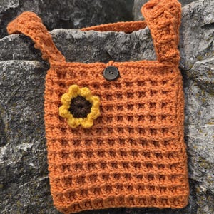 May include: An orange crochet bag with a waffle stitch pattern. The bag features a sunflower applique and a button closure. The bag has two straps and is made of a soft, textured yarn.