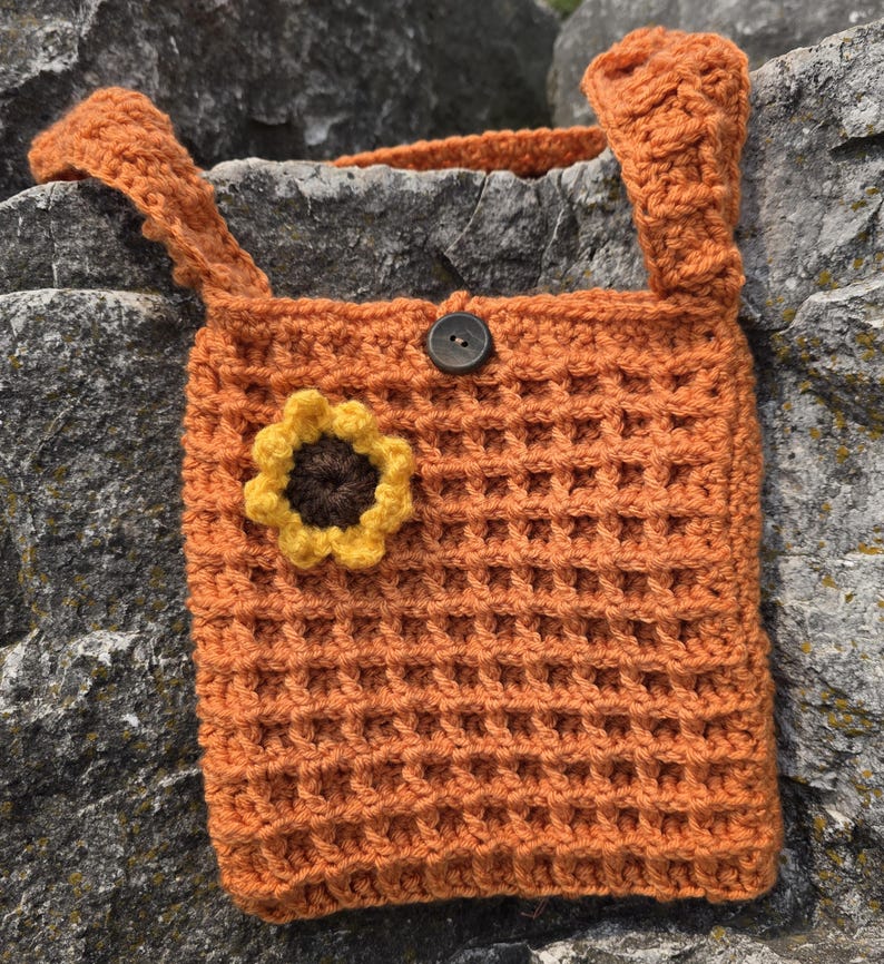 May include: An orange crochet bag with a waffle stitch pattern. The bag features a sunflower applique and a button closure. The bag has two straps and is made of a soft, textured yarn.