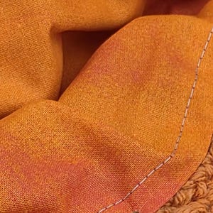 May include: Close-up of a folded orange fabric with a gradient of red and orange hues. The fabric is stitched with a white thread along the edge. A textured, crocheted orange fabric is visible on the right side.