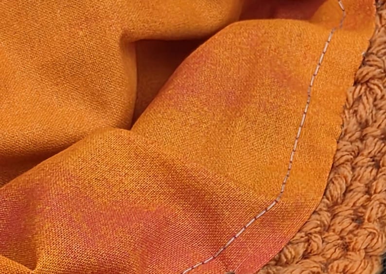 May include: Close-up of a folded orange fabric with a gradient of red and orange hues. The fabric is stitched with a white thread along the edge. A textured, crocheted orange fabric is visible on the right side.
