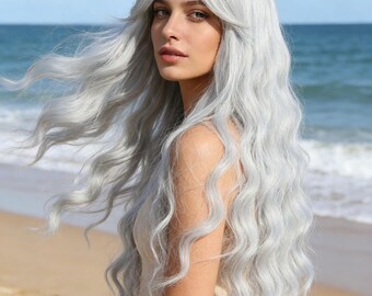 May include: A woman with long, wavy, silver-gray hair stands on a beach. The hair has a natural wave and a slight fringe. She wears a light-colored dress. The background shows the ocean and a blue sky.