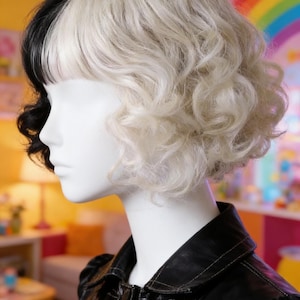 May include: A mannequin head with a two-toned wig, black on the left and white on the right, styled in short, curly layers. The mannequin wears a black leather jacket with white stitching. The background features a rainbow and colorful decor.