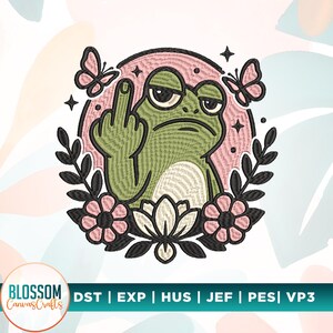 May include: An embroidery design featuring a green frog with a grumpy expression giving the middle finger, set against a pink circular background. The frog is framed by black leafy branches, pink flowers, and two butterflies, with the text Blossom CanvasCrafts and file formats DST, EXP, HUS, JEF, PES, VP3.