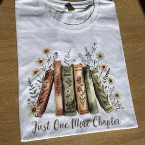 Könnte beinhalten: Weißes T-Shirt mit einem Design aus fünf Büchern, floralen Elementen und dem Text "Just One More Chapter". Die Bücher sind in Braun-, Grün- und Beigetönen gehalten, mit Gänseblümchen- und Blattillustrationen.