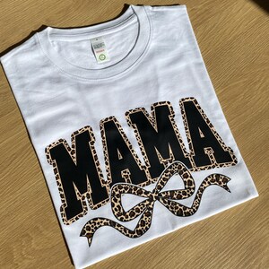 Könnte beinhalten: Weißes T-Shirt mit dem Wort "MAMA" in großen schwarzen Buchstaben mit Leopardenmuster-Umrandung. Unter dem Wort befindet sich eine Schleife mit Leopardenmuster. Das Shirt liegt auf einer hellbraunen Holzoberfläche.