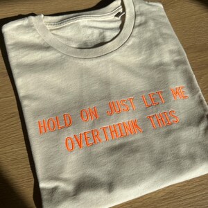 Pode incluir: Camiseta creme com o texto bordado em laranja "HOLD ON JUST LET ME OVERTHINK THIS". A camiseta está dobrada sobre uma superfície de madeira, com sombras criadas pela luz solar.