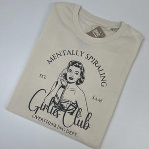 Könnte beinhalten: Beiges T-Shirt mit schwarzer Grafik einer Frau am Telefon und dem Text "MENTALLY SPIRALING, Girlies Club, OVERTHINKING DEPT." Das Shirt hat einen Rundhalsausschnitt und ein Etikett. Es liegt gefaltet auf einer weißen Oberfläche.