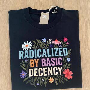 Può includere: Felpa nera con la scritta "RADICALIZED BY BASIC DECENCY" in lettere colorate, circondata da illustrazioni floreali. L'etichetta recita "Organic Cotton, Coton Bio".