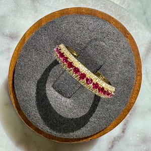 May include: A gold band ring featuring a central row of vibrant round-cut pink stones flanked by two thinner rows of small, clear white stones. The ring is designed with a polished metallic finish and a consistent, symmetrical arrangement of gemstones.