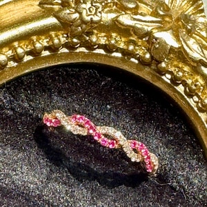 May include: A twisted band ring featuring alternating strands of sparkling clear stones and vibrant pink stones. The ring is set against a dark, textured surface and framed by a decorative, ornate gold-colored border with floral detailing.