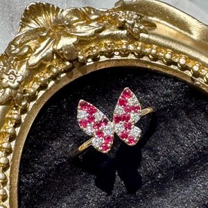 May include: A gold ring featuring a butterfly-shaped centerpiece adorned with a pattern of small, round, sparkling red and clear white stones. The butterfly design is positioned centrally, showcasing a vibrant contrast between the colored gems.