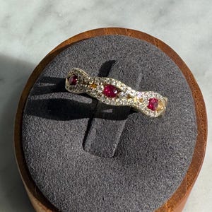 May include: A gold band ring featuring a wavy, textured design set with three round, vibrant red rubies and numerous small, clear pavé-set diamonds along the surface, creating a sparkling, multi-stone appearance.