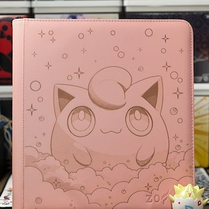 May include: A pink card binder featuring a cartoon character design. The binder has a zipper closure and a stitched border. The design includes a character in a cloud with stars and bubbles. A small figurine is in the foreground.