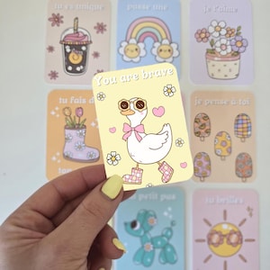 Peut inclure: Plusieurs cartes illustrées avec des fonds pastel. L'une d'elles représente une oie portant des lunettes de soleil et des bottes, avec le texte « You are brave ». D'autres cartes montrent un arc-en-ciel, des fleurs et des glaces.