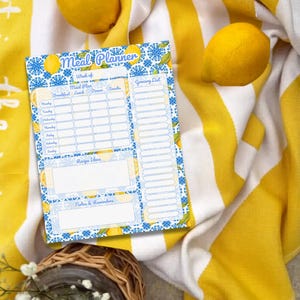 May include: A meal planner with a blue and white lemon design, featuring sections for meal planning, a grocery list, and notes. The planner is placed on a yellow and white striped towel, with fresh lemons scattered around. A basket with white flowers is in the foreground.