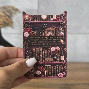 May include: A rectangular card featuring a dark, magical bookshelf illustration with a black cat, pink roses, and vintage books. The text reads: ALWAYS THROW SPILT SALT OVER YOUR LEFT SHOULDER, KEEP ROSEMARY BY YOUR GARDEN GATE, PLANT LAVENDER FOR LUCK, AND FALL IN LOVE WHENEVER YOU CAN.