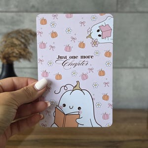 Op de afbeelding: Een rechthoekige kaart met een lichtpaarse achtergrond met een herhalend patroon van kleine oranje pompoenen, witte madeliefjes en roze strikjes. De tekst "Just one more Chapter" staat in het midden, vergezeld door twee witte spookillustraties.