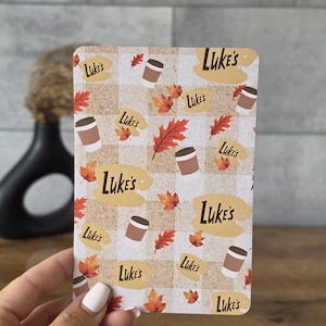 Può includere: Una carta rettangolare con un motivo a quadri beige e bianco decorata con una stampa ripetuta di tazze da caffè marroni, foglie autunnali arancioni e rosse e il testo Luke's scritto in corsivo marrone su forme ovali color sabbia.