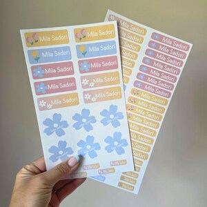 Peut inclure: Deux feuilles d'étiquettes adhésives rectangulaires et en forme de fleur dans des tons pastel jaune, bleu et rose. Chaque étiquette porte le texte "Mila Sadori" en blanc, accompagné de petites illustrations florales disposées en colonnes.