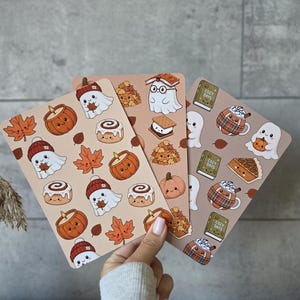 Op de afbeelding: Drie rechthoekige stickervellen met herfstachtige illustraties. De ontwerpen bevatten geesten met mutsen, lachende pompoenen, kaneelbroodjes, esdoornbladeren, s'mores, boeken met het label Cozy Days en mokken met ruitpatronen in warme bruintinten.