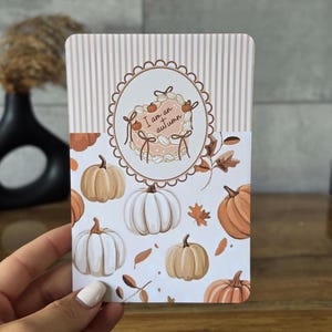 Op de afbeelding: Een rechthoekige kaart met een bovenkant met beige en witte strepen en een witte onderkant versierd met diverse oranje, witte en lichtbruine pompoenen en bladeren. Een centrale ovale omlijsting bevat een taart met de tekst I am an autumn.