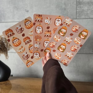 Peut inclure: Trois feuilles d'autocollants rectangulaires présentant un motif répétitif d'illustrations comprenant des hérissons dans des citrouilles, des tasses de chocolat chaud avec de la crème fouettée, des roulés à la cannelle et des feuilles d'automne sur un fond beige clair.