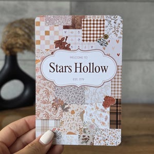May include: A rectangular card featuring a collage of various patterns in shades of brown, beige, and cream, including gingham, floral, and checkered prints. Centered is a white label with the text "WELCOME TO Stars Hollow EST. 1779".