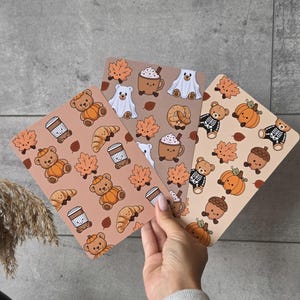 May include: Three rectangular cards featuring repeating patterns of autumn-themed illustrations. The designs include brown teddy bears wearing pumpkin costumes, ghost-clad bears, skeleton-clad bears, orange maple leaves, coffee cups, and croissants.