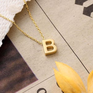 May include: A gold-toned necklace featuring a capital letter B pendant suspended from a delicate matching chain. The pendant has a smooth, polished finish and a block-style font, reflecting light across its surface while hanging from a thin link chain.