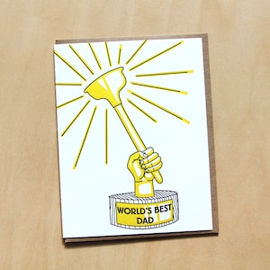 Op de afbeelding: Een gele en zwarte kaart met een cartoonillustratie van een hand die een zuiger trofee vasthoudt. De trofee heeft een gele basis met de woorden "World's Best Dad" erop.