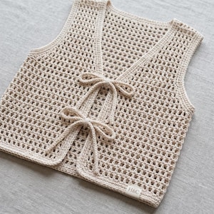 Op de afbeelding: Een beige, open geweven gehaakt vest met een V-hals en striksluitingen. Het vest heeft een delicaat, luchtig ontwerp en een klein label met de letters "H&C". Het handgemaakte kledingstuk is waarschijnlijk voor layering.