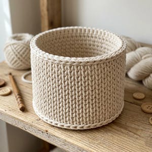 May include: A beige, cylindrical crochet basket. The basket is made of thick yarn and has a textured appearance. The basket is sitting on a wooden surface, with a ball of yarn and other craft supplies in the background.