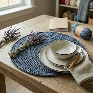 Peut inclure: Mise en place rustique sur une table en bois. Un set de table bleu crocheté, des assiettes et un bol en céramique mouchetée, des couverts et une serviette en lin. De la lavande séchée et une pelote de laine complètent la scène.