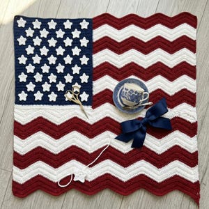 May include: A handmade crochet blanket in the style of the American flag, featuring red and white chevron stripes and a blue square with white star appliqués. A blue and white teacup and saucer, a navy blue bow, and a pair of scissors are on the blanket.