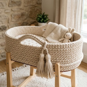 May include: A beige crochet baby bassinet with a floral patterned interior, a matching blanket, and a stuffed bunny. The bassinet has a handle and decorative tassels, and is supported by a light wood stand.