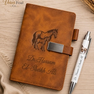 May include: A tan leather-like notebook with a metal clasp, featuring an engraved horse and foal design and the text Dr. Hossam El-Sheikh Ali. Beside it lies a white ballpoint pen with a matching horse graphic and the same engraved name.