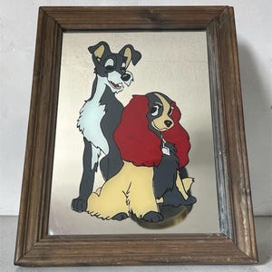 Peut inclure: Miroir encadré avec une image de dessin animé de deux chiens, l'un noir et blanc, l'autre beige avec des cheveux rouges. Le cadre est en bois marron. Le miroir a une base dorée.