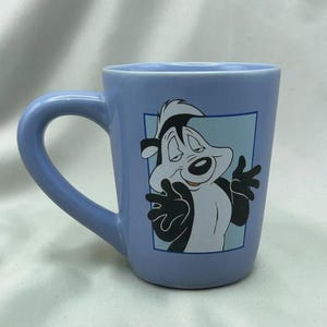 Puede incluir: Taza de cerámica azul claro con un personaje de dibujos animados de mofeta. La mofeta tiene pelaje blanco y negro y está sobre un fondo azul claro. La taza tiene un asa curvada.